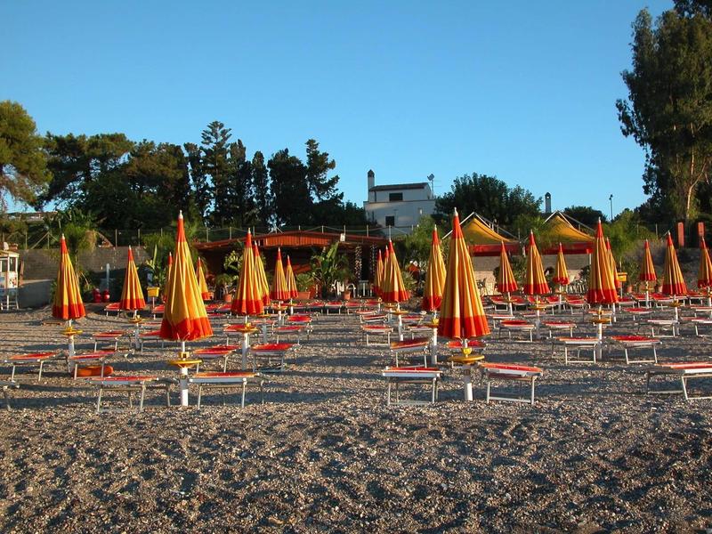 Many closed beach umbrellas and lounge chairs on a pebble beach under a clear sky.