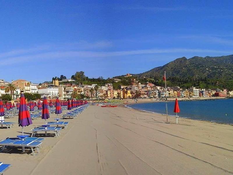 Wide sandy beach with sun loungers, red umbrellas, and clear blue sky by the sea.