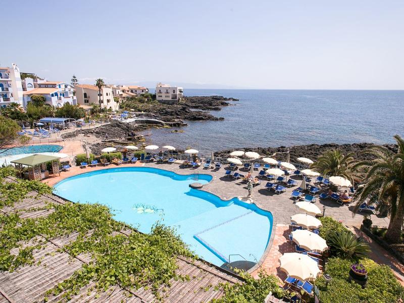 Hotel pool with loungers and palm trees next to rocky coast and sea under clear sky.