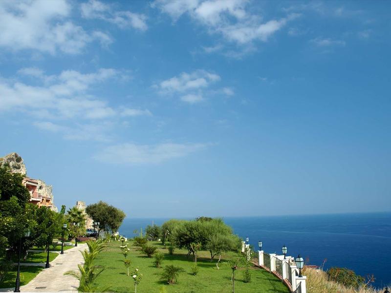Grüner Park mit Schritten, Bäumen und Bänken neben blauem Meer und blauem Himmel mit Wolken.