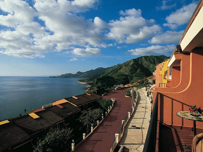 Küste mit blauen Meer, grün bewachsenen Hügeln, bunten Häusern und bewölktem Himmel.