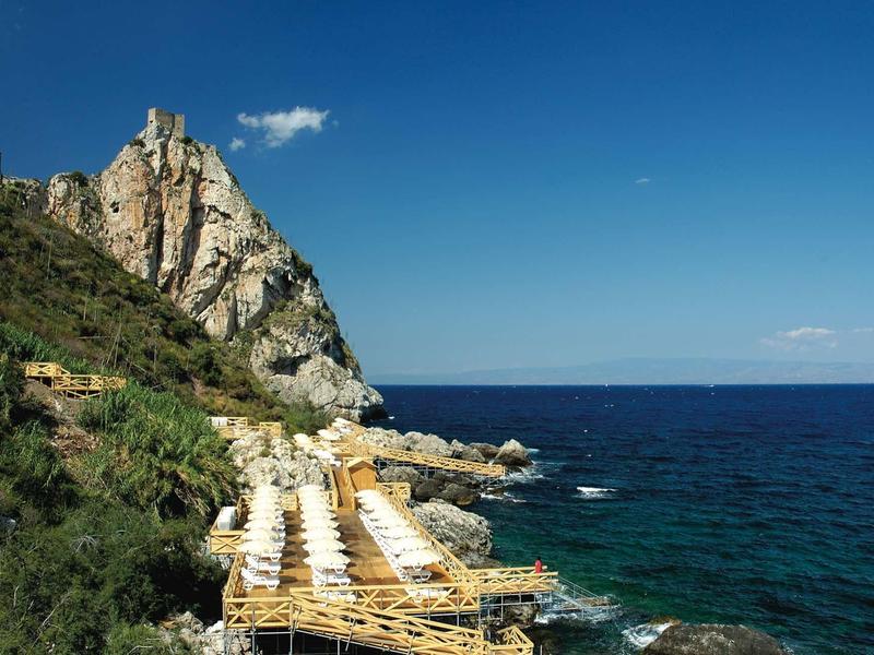 Klippen mit grüner Vegetation und Holzstegen mit weißen Liegestühlen am blauen Meer unter klarem Himmel.