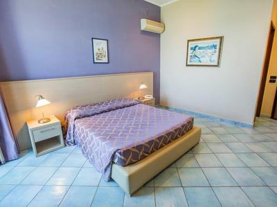 Světle zařízený hotelový pokoj s velkou postelí, nočními lampami a dlaždicovou podlahou.