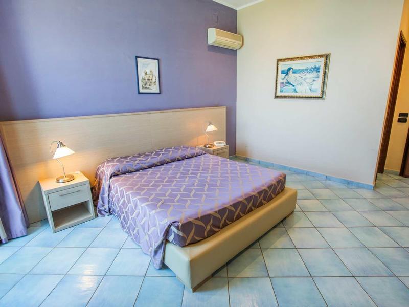 Světle zařízený hotelový pokoj s velkou postelí, nočními lampami a dlaždicovou podlahou.