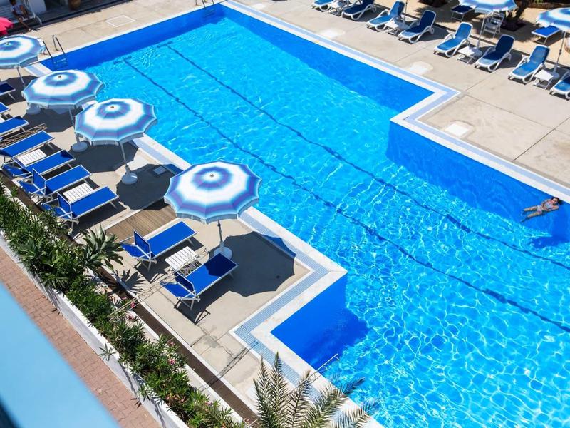 Pohled shora na velký modrý bazén s modrými lehátky a bílými slunečníky.