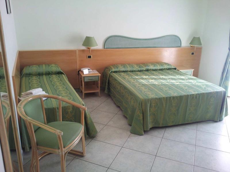 Camera d'hotel con due letti, copriletti verdi e una sedia accanto al comodino.