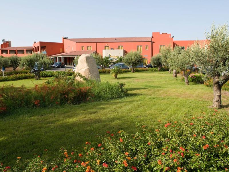 Gran edificio de hotel rojo detrás de un jardín cuidado con césped y olivos.