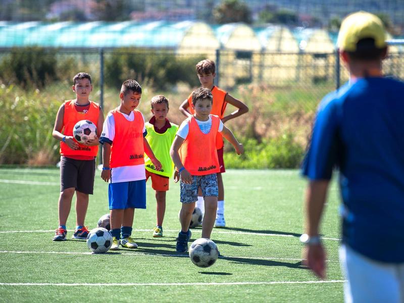 Niños en chalecos jugando al fútbol en un campo mientras un entrenador los observa.