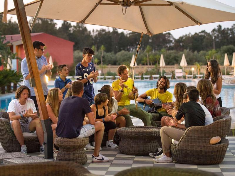 Grupo de personas relajándose en el área exterior de un hotel bajo una sombrilla junto a la piscina.
