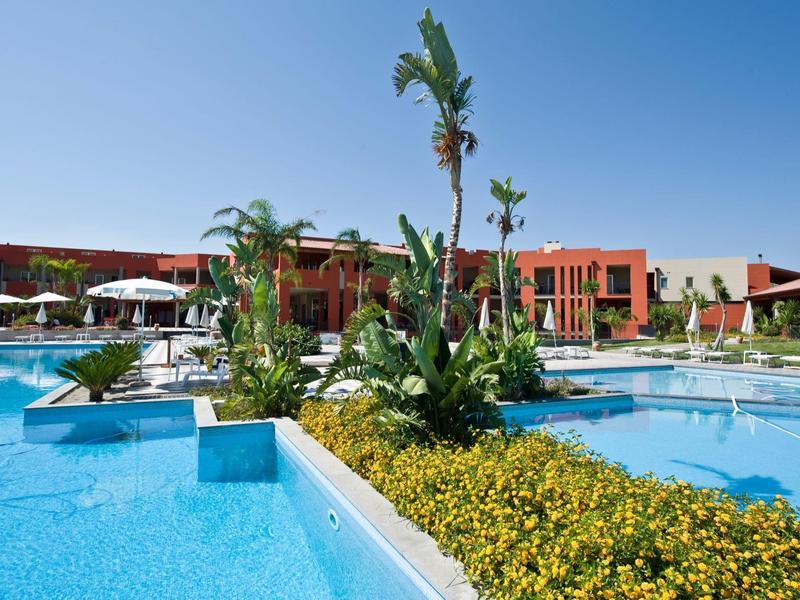 Gran piscina de hotel con palmeras y edificios rojos al fondo bajo un cielo soleado.