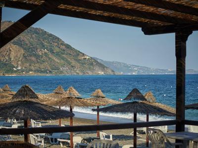 View from a covered terrace overlooking a beach promenade with straw umbrellas and the sea beyond.