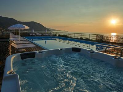 Hot tub on terrace overlooking a pool, mountains, and sunset over the sea.