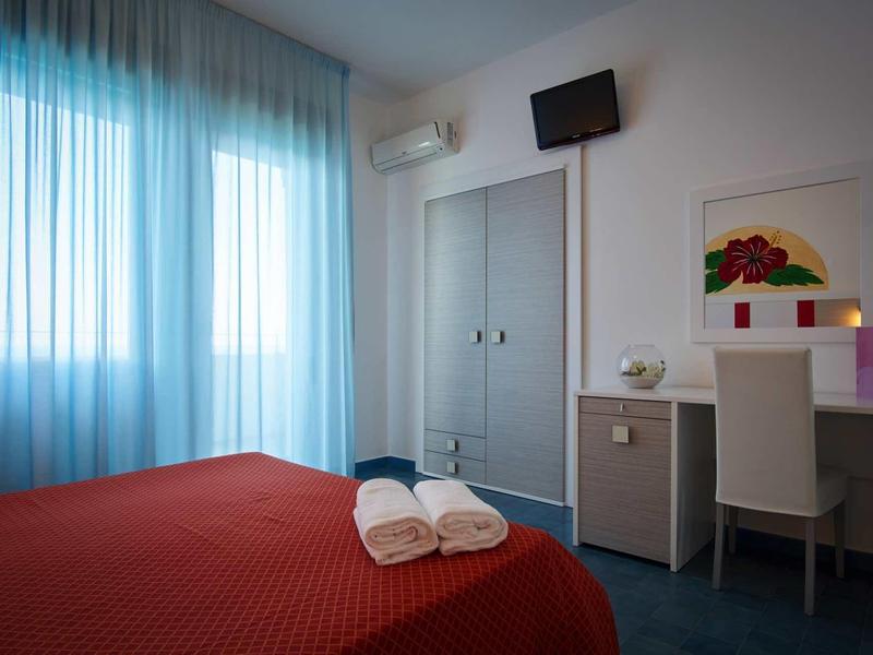Simple hotel room with red bed, white furniture, and blue curtains.