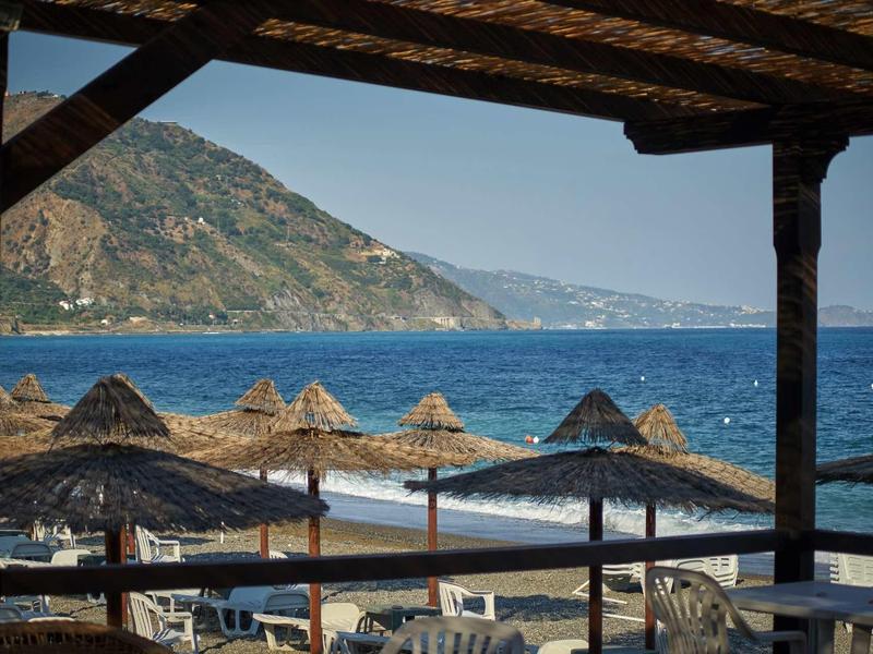 View from a covered terrace overlooking a beach promenade with straw umbrellas and the sea beyond.