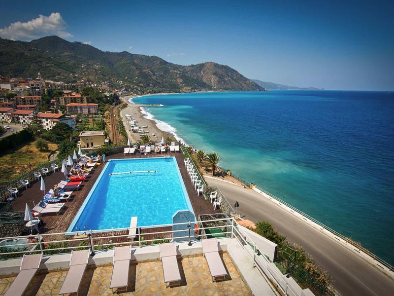 View of a pool and beach along the coast with mountains in the background under clear sky.