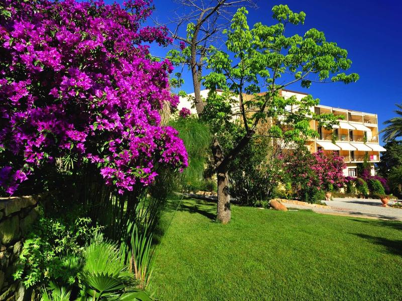 Hotelová budova za barevnou zahradou s zeleným trávníkem a modrou oblohou.