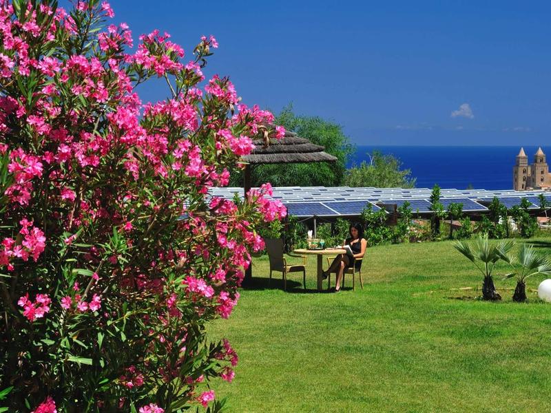 Kvetoucí keř oleandru vedle zelené zahrady s výhledem na moře a venkovními stoly.