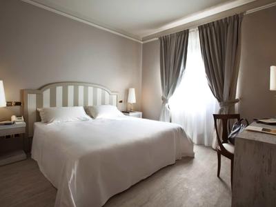 Světle hotelový pokoj s manželskou postelí, nočními stolky a závěsy u okna.