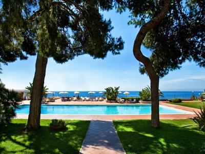 Blick auf einen Pool mit Liegestühlen, umgeben von Bäumen und blauem Himmel am Meer im Hintergrund.