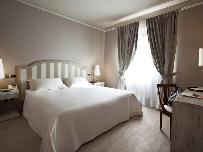 Světle hotelový pokoj s manželskou postelí, nočními stolky a závěsy u okna.