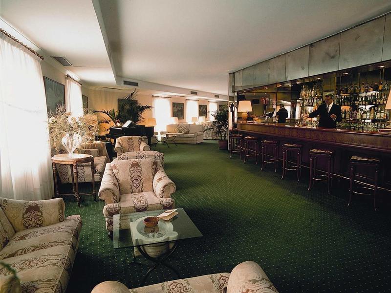 Elegantní hotelový salonek se zeleným kobercem, pohodlnými křesly a dlouhým barem se zrcadlovou stěnou.