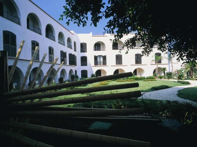 Hotelové nádvoří s bílou fasádou, arkádami a upravenou zahradou pod modrou oblohou.