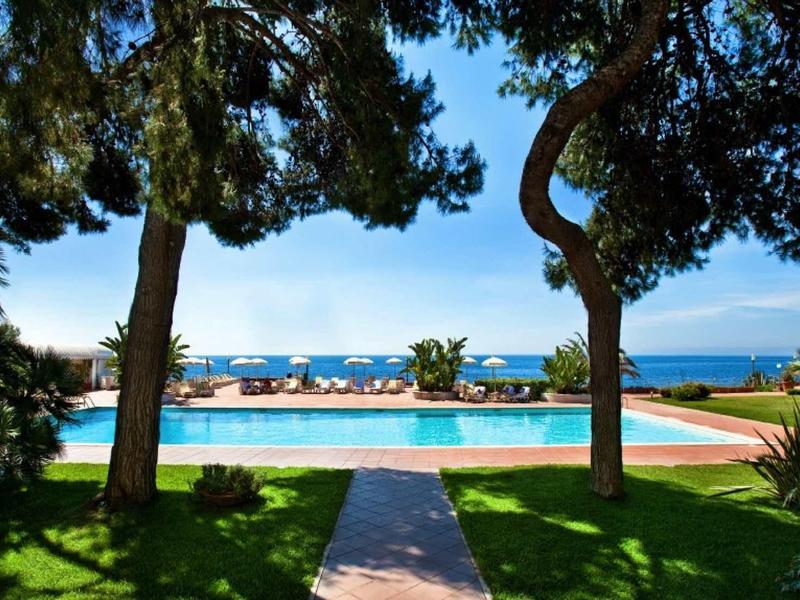 Pohled na bazén s lehátky, obklopený stromy a s mořem v pozadí.