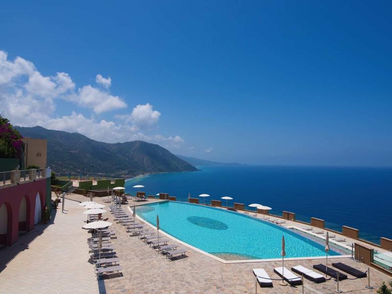 Infinity zwembad met ligstoelen met uitzicht op zee en bergen bij heldere lucht.