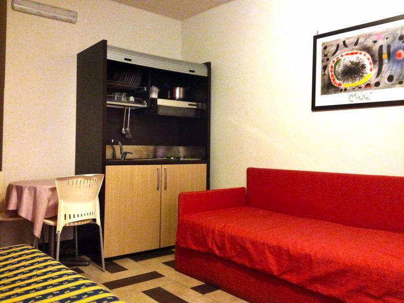 Small hotel room with red sofa, desk, and open kitchenette with cabinets.