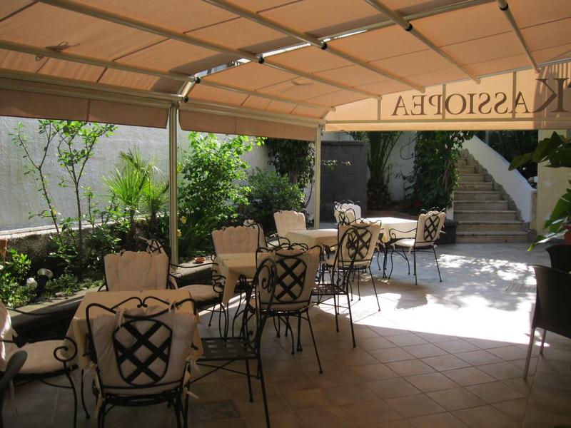 Covered outdoor area with tables and chairs, surrounded by plants and sunlight.