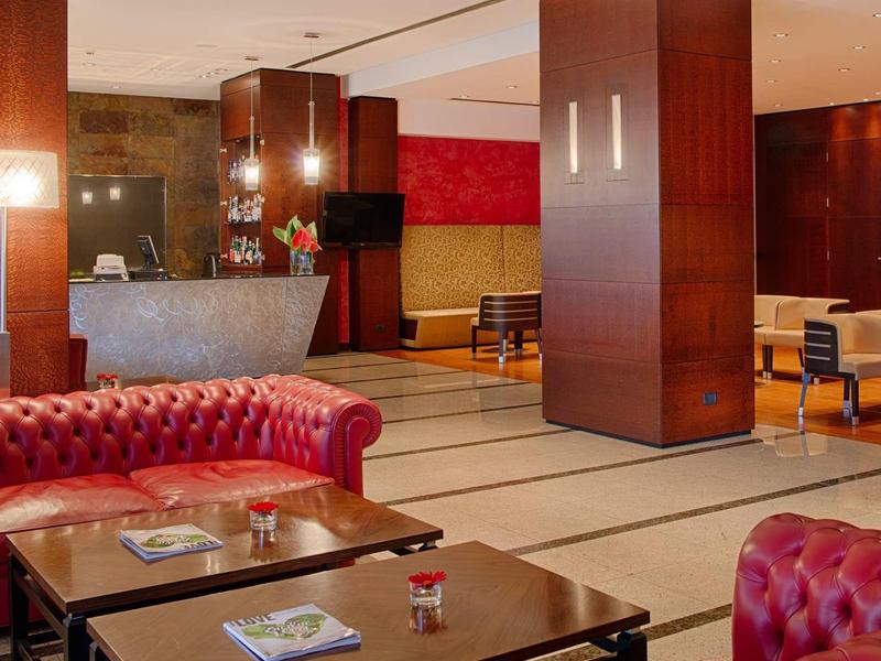 Hall dell'hotel con divano Chesterfield rosso, tavoli e reception moderna sullo sfondo.