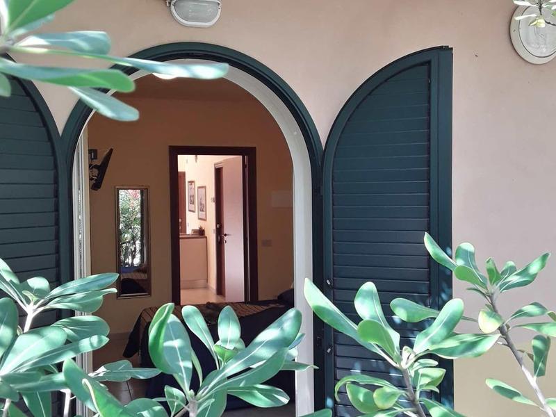 Vista attraverso foglie verdi verso un ingresso con persiane blu aperte e interno caldo.