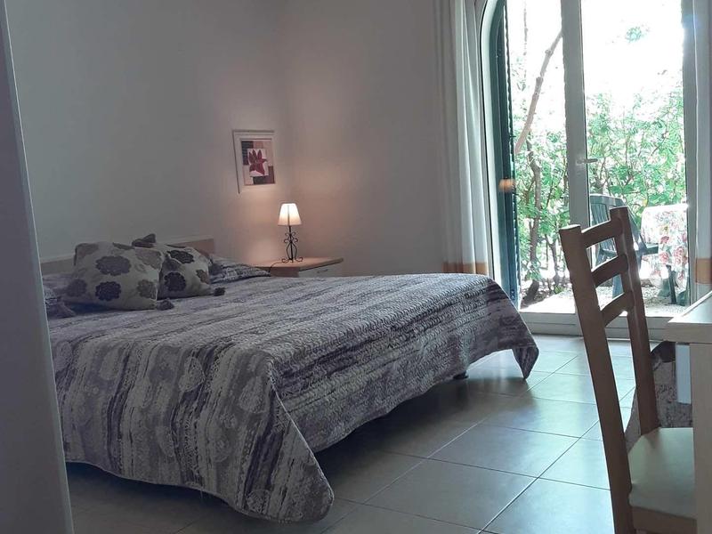 Camera semplice con letto matrimoniale, lampada da comodino e grande finestra ad arco con vista sul verde.