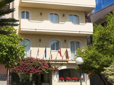 Mehrstöckiges weißes Gebäude mit Balkonen, grünen Bäumen und roter Blumentracht im vorderen Bereich.