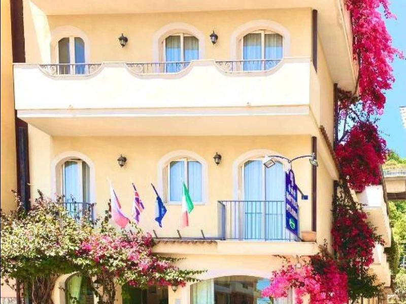 Hotel di tre piani con balconi e piante fiorite sulla facciata in una strada soleggiata.