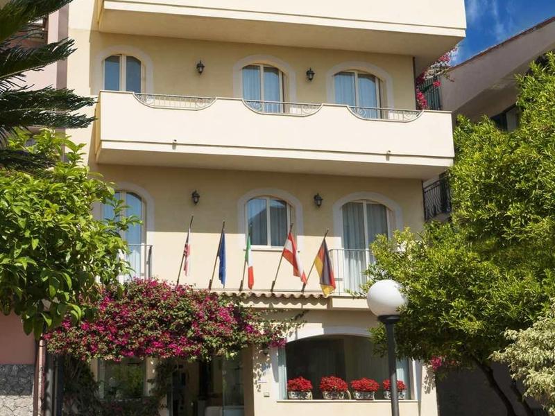 Hotel di tre piani con facciate bianche, balconi e zona pranzo esterna sotto un cielo azzurro.