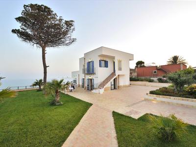 Weißes Haus mit blauen Fensterläden, umgeben von Garten und großem Baum am Meer.