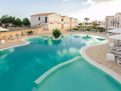 Großer Pool mit klar blauem Wasser, Sonnenliegen und weißen Häusern im Hintergrund bei Tageslicht.
