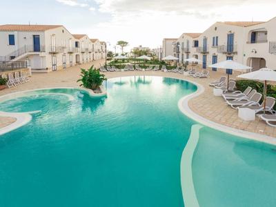 Großer, geschwungener Pool mit weißen Liegen umgeben von weiß-blauen Ferienhäusern bei hellem Himmel.