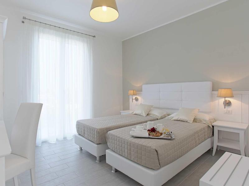 Helles Schlafzimmer mit zwei Einzelbetten, weißem Mobiliar, grauer Wand und Tageslicht durch Fenster.