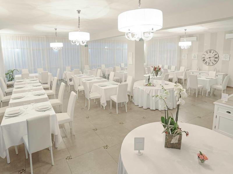 Heller, großzügiger Speisesaal mit weißen Tischen, Stühlen und modernen Kronleuchtern.