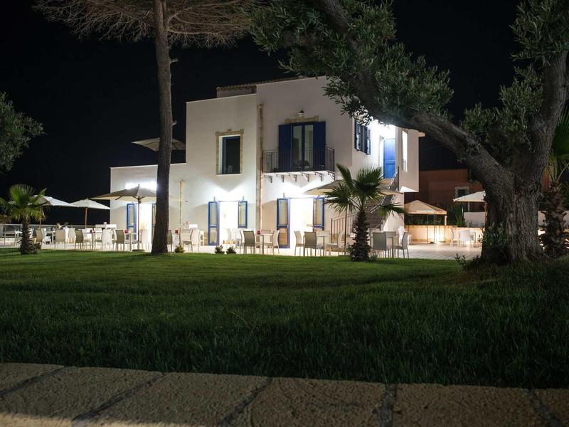 Ein beleuchtetes weißes Haus mit Balkon und Terrasse umgeben von Bäumen bei Nacht.