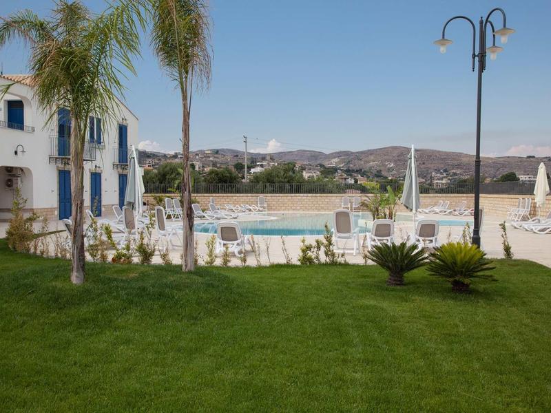 Grüner Rasen mit Palmen vor einem weißen Gebäude, Schwimmbecken und Bergen im Hintergrund bei klarem Himmel.