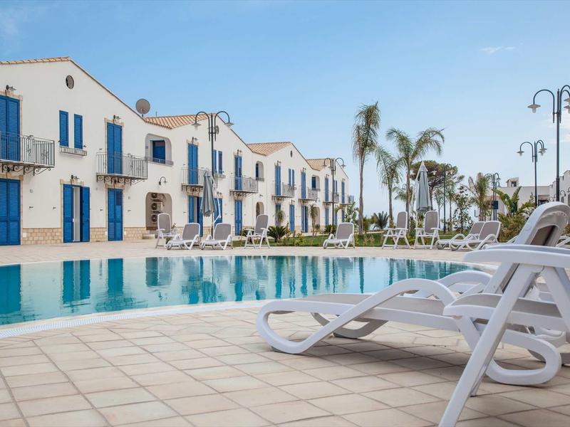 Großer Pool vor weißen Häusern mit blauen Türen und Palmen bei klarem Himmel.