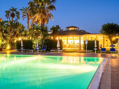 Piscina illuminata davanti a un edificio dell'hotel con palme al crepuscolo.