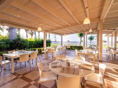 Ristorante luminoso e aperto con tetto in legno, tavoli e sedie bianchi, vista su palme e mare.