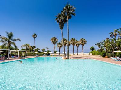 Grande piscina con acqua blu chiara circondata da palme e ombrelloni all'hotel.
