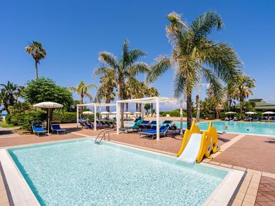 Area piscina dell'hotel con piscina per bambini, scivolo e palme sotto un cielo sereno.