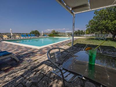 Sonniger Poolbereich mit Liegestühlen und Getränk auf Glastisch, grüne Umgebung und blauer Himmel.