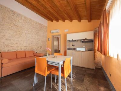 Helles Zimmer mit Esstisch, orangenen Stühlen, Sofa und Küchenzeile unter Holzbalkendecke.